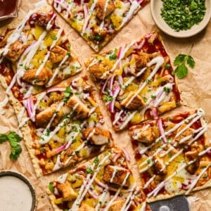 Lavash pizza slices topped with grilled chicken, onions, and herbs, drizzled with white sauce. Arranged on parchment with small bowls of herbs and sauce beside a pizza cutter.