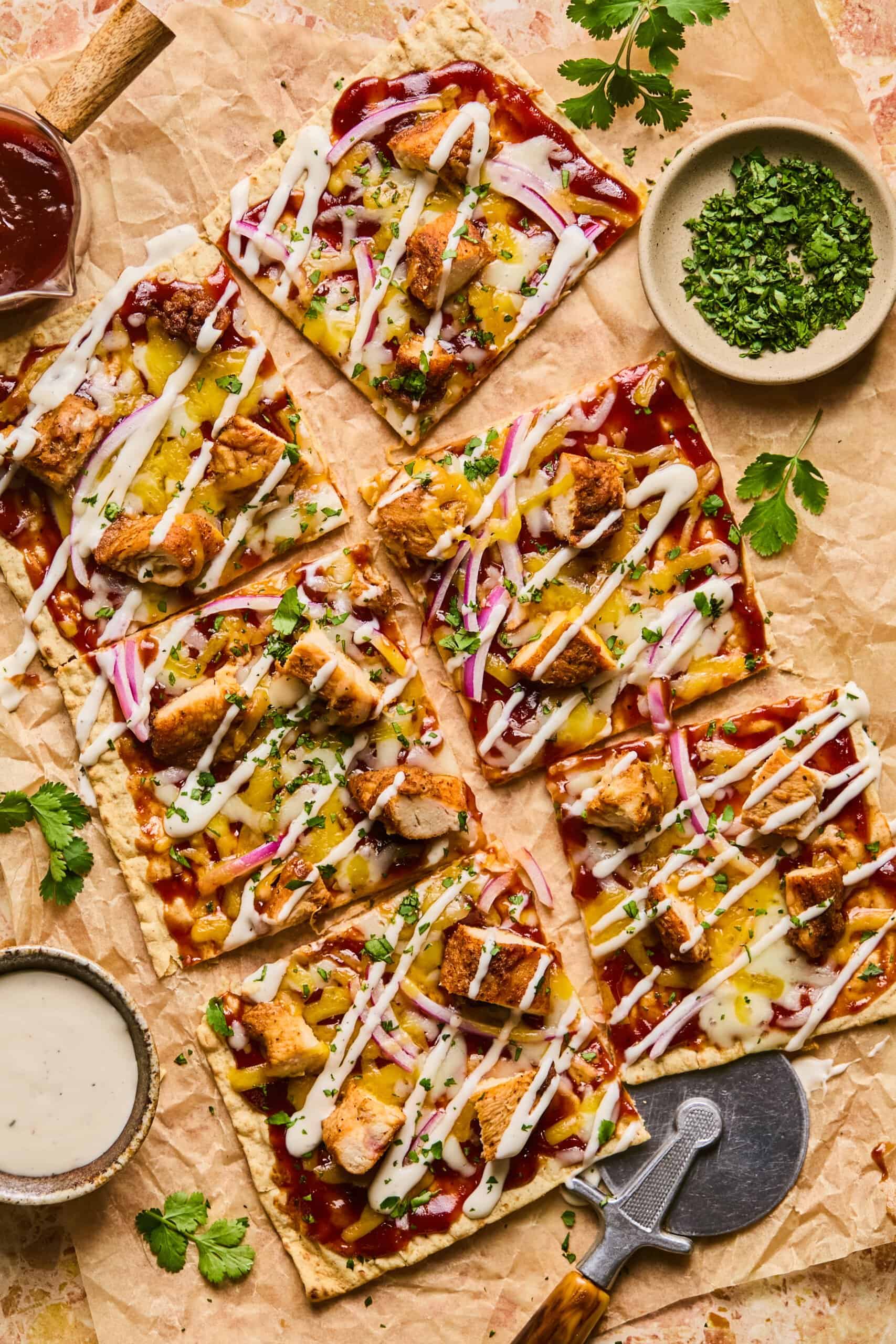 Lavash pizza slices topped with grilled chicken, onions, and herbs, drizzled with white sauce. Arranged on parchment with small bowls of herbs and sauce beside a pizza cutter.