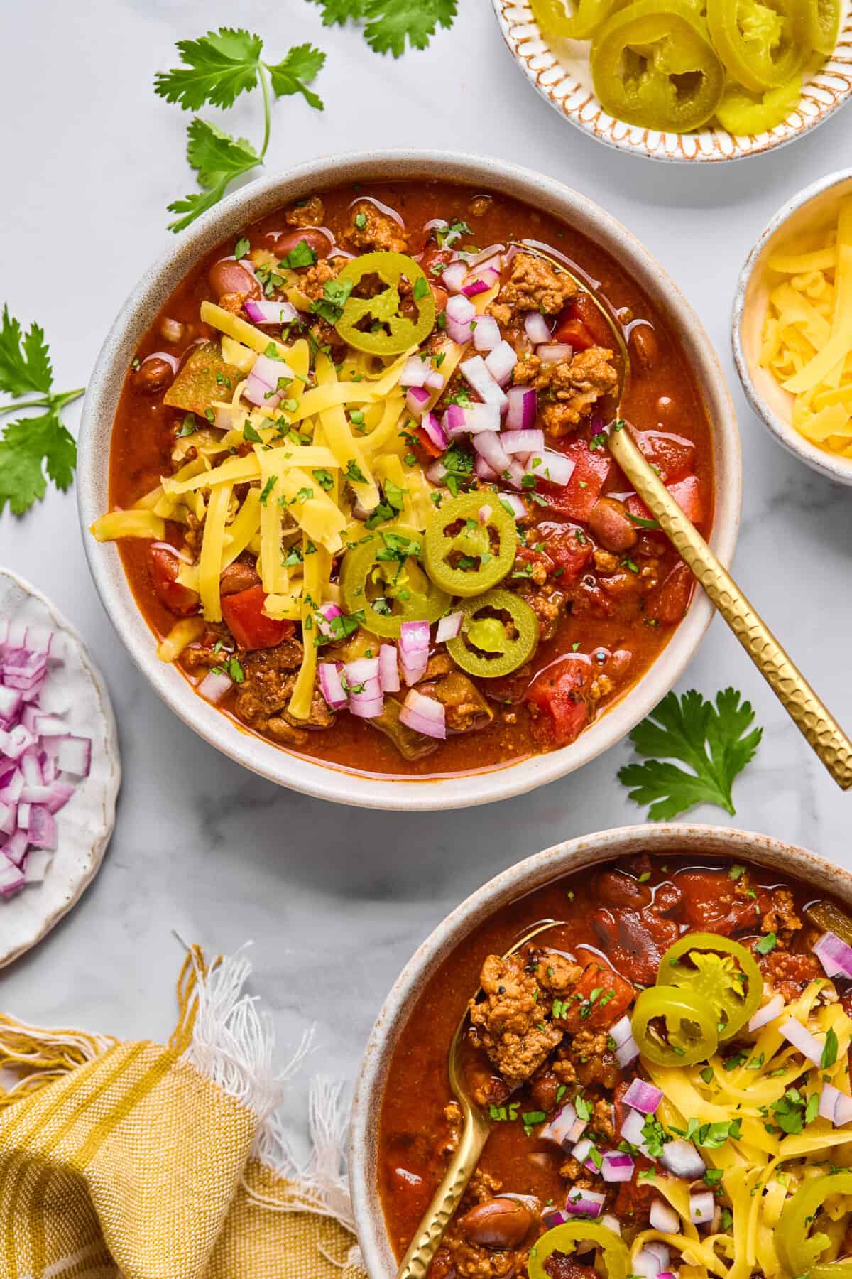 Two bowls of macro friendly chili topped with shredded cheese, sliced jalapeรฑos, and chopped red onions, surrounded by small bowls of extra toppings and fresh cilantro on a light surface.