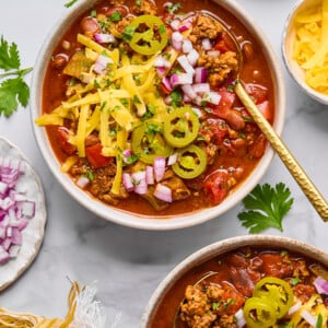Two bowls of macro friendly chili topped with shredded cheese, sliced jalapeรฑos, and chopped red onions, surrounded by small bowls of extra toppings and fresh cilantro on a light surface.