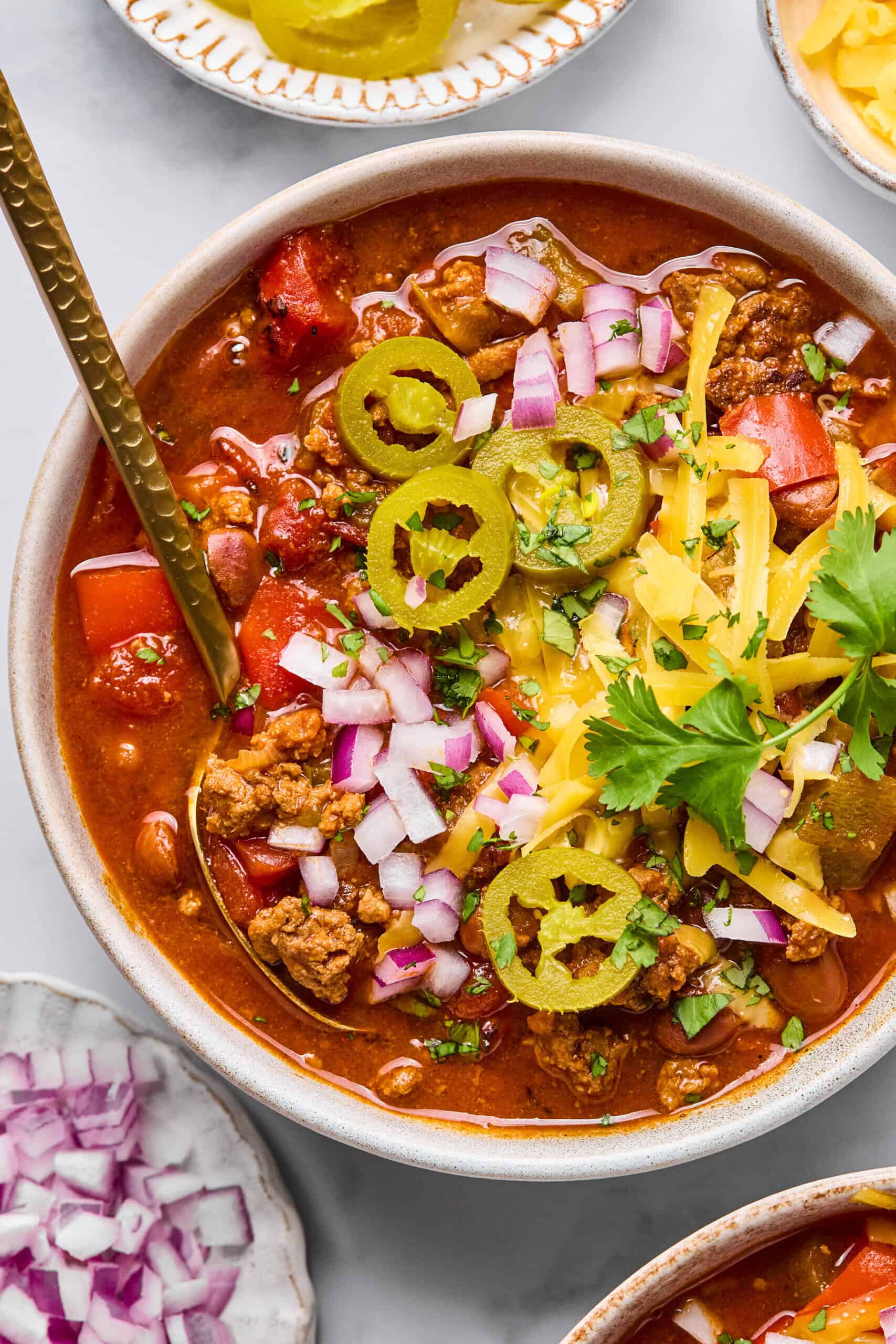A bowl of macro friendly chili topped with sliced jalapeños, shredded cheese, chopped red onions, and fresh cilantro, with a spoon inside. Surrounding dishes offer extra toppings for customization.