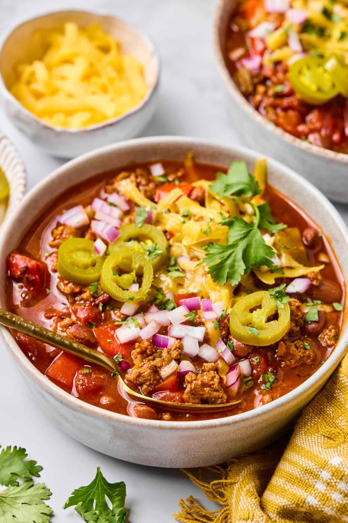 A bowl of macro friendly chili topped with shredded cheese, chopped red onions, sliced jalapeรฑos, and fresh cilantro, with another bowl and a dish of cheese in the background. A yellow napkin and cilantro leaves are nearby.