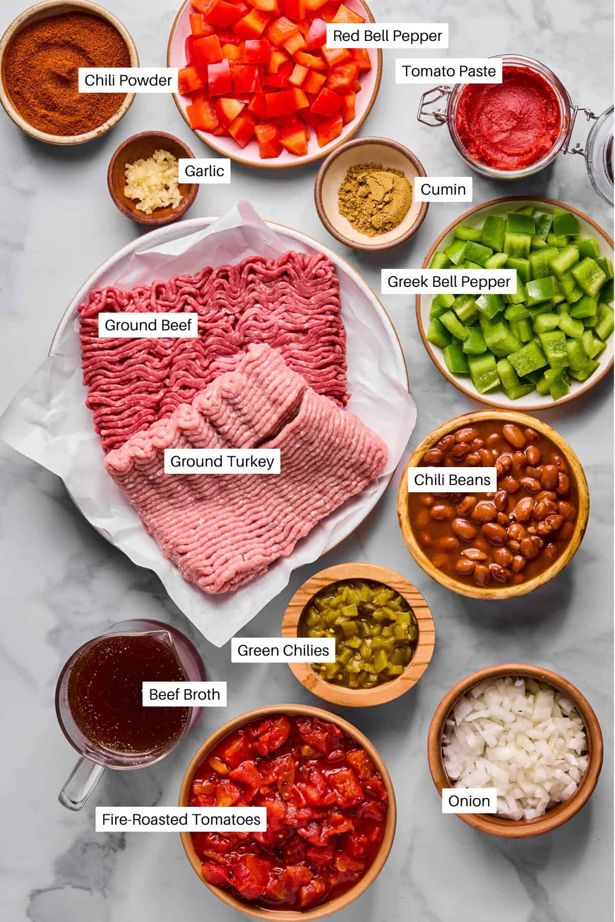 A flat lay of labeled macro friendly chili ingredients: chili powder, red bell pepper, tomato paste, garlic, cumin, green bell pepper, ground beef, ground turkey, chili beans, green chiles, beef broth, fire-roasted tomatoes, and chopped onion.