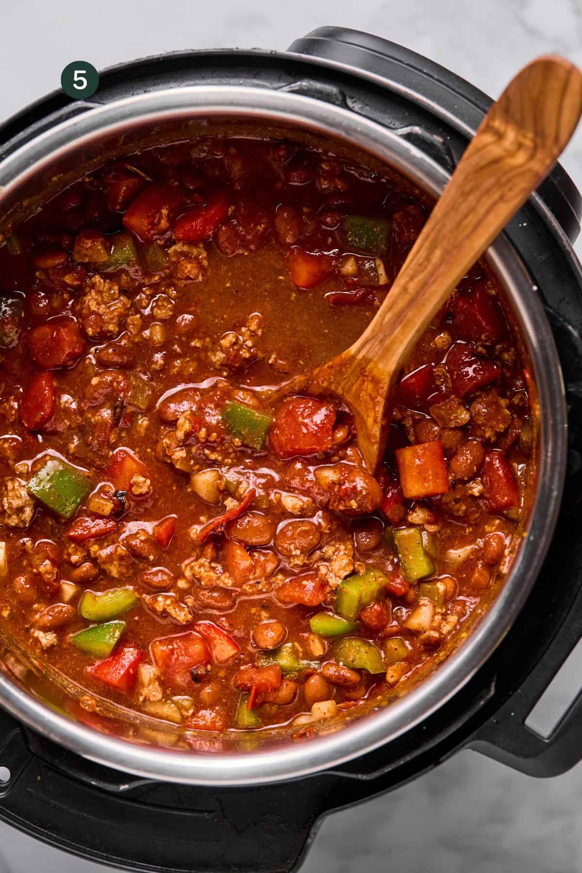 A pot of macro friendly chili with ground meat, beans, tomatoes, green peppers, and carrots is being stirred with a wooden spoon inside a pressure cooker.