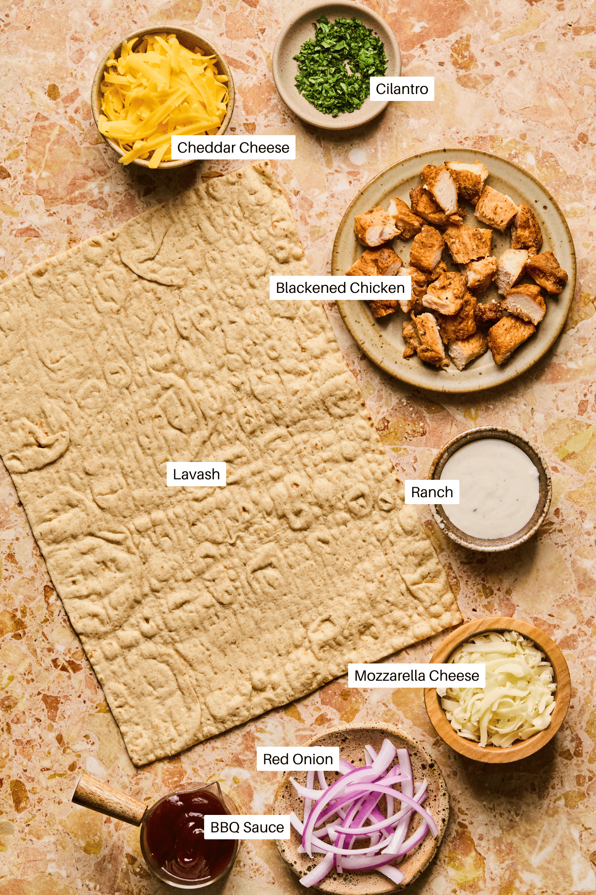 Ingredients for Lavash Pizza are laid out on a speckled countertop. Included are lavash bread, cheddar and mozzarella cheese, blackened chicken, red onion, BBQ sauce, cilantro, and ranch dressing—each meticulously placed in separate bowls or plates.