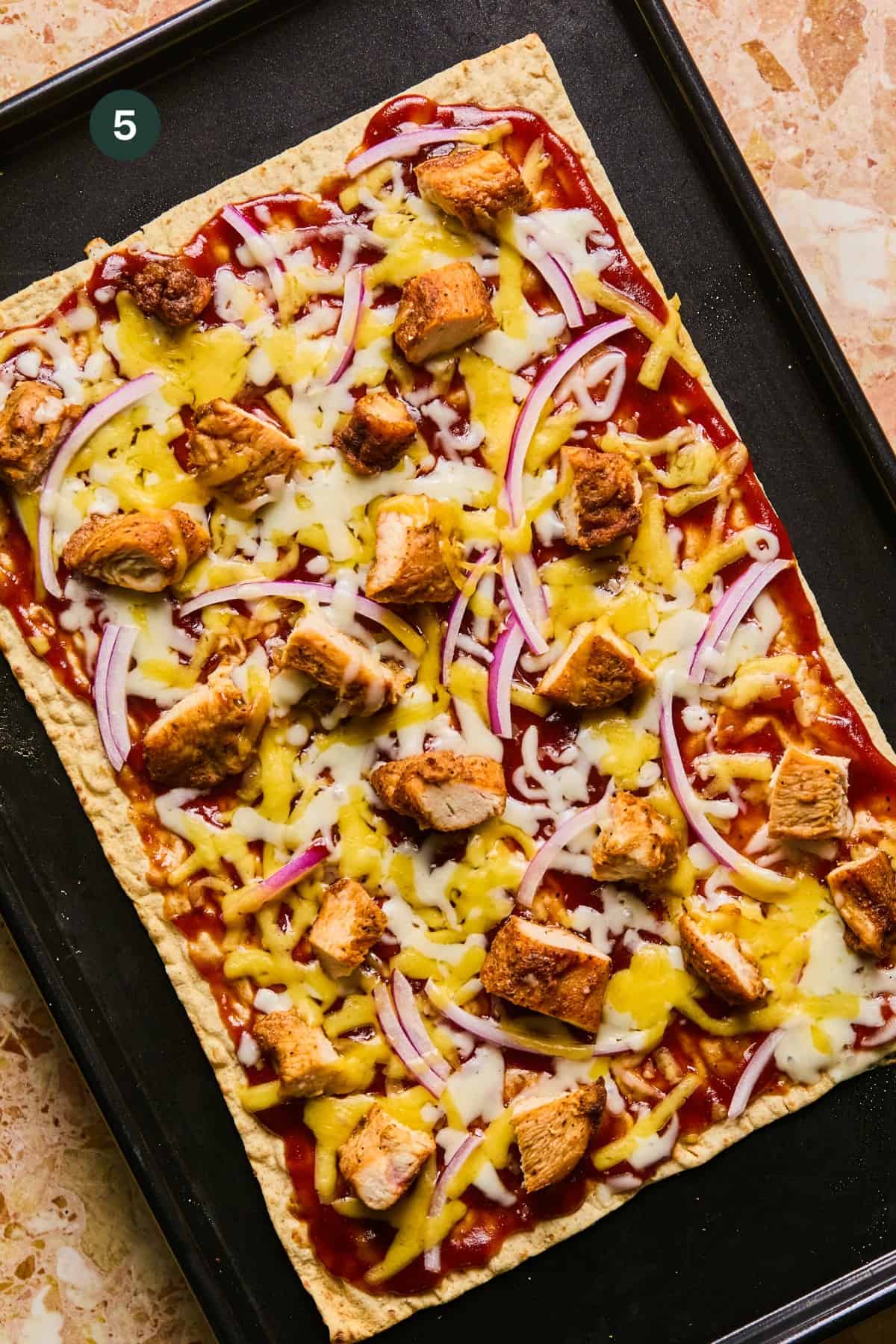 A rectangular lavash pizza topped with barbecue sauce, grilled chicken chunks, sliced red onions, and melted cheese rests on a black baking tray. The background is a beige stone surface.