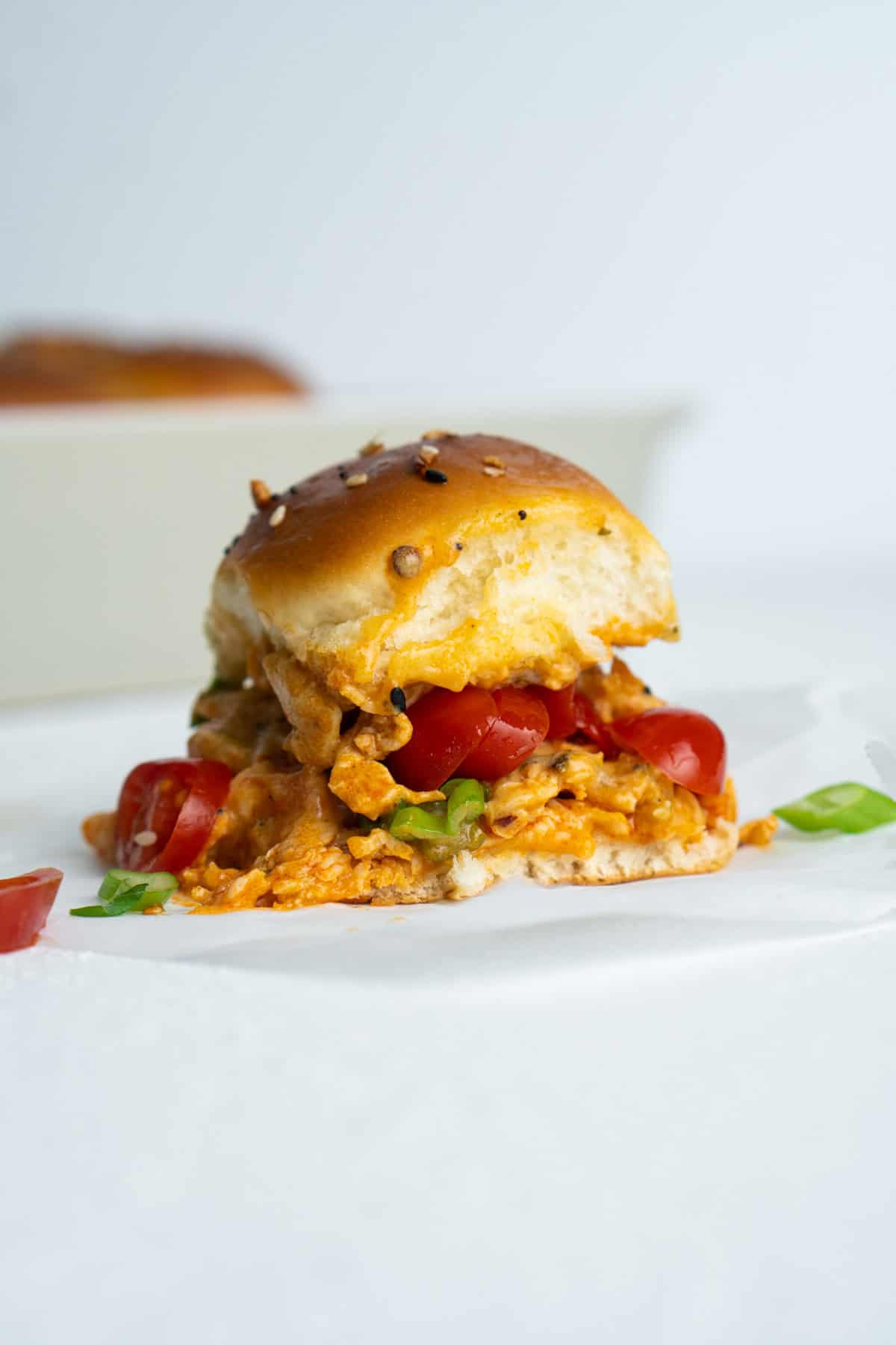 A buffalo chicken slider with a fluffy bun sits on a white surface. The filling features shredded chicken in a spicy sauce, sliced cherry tomatoes, and chopped green onions. A white baking dish is blurred in the background.