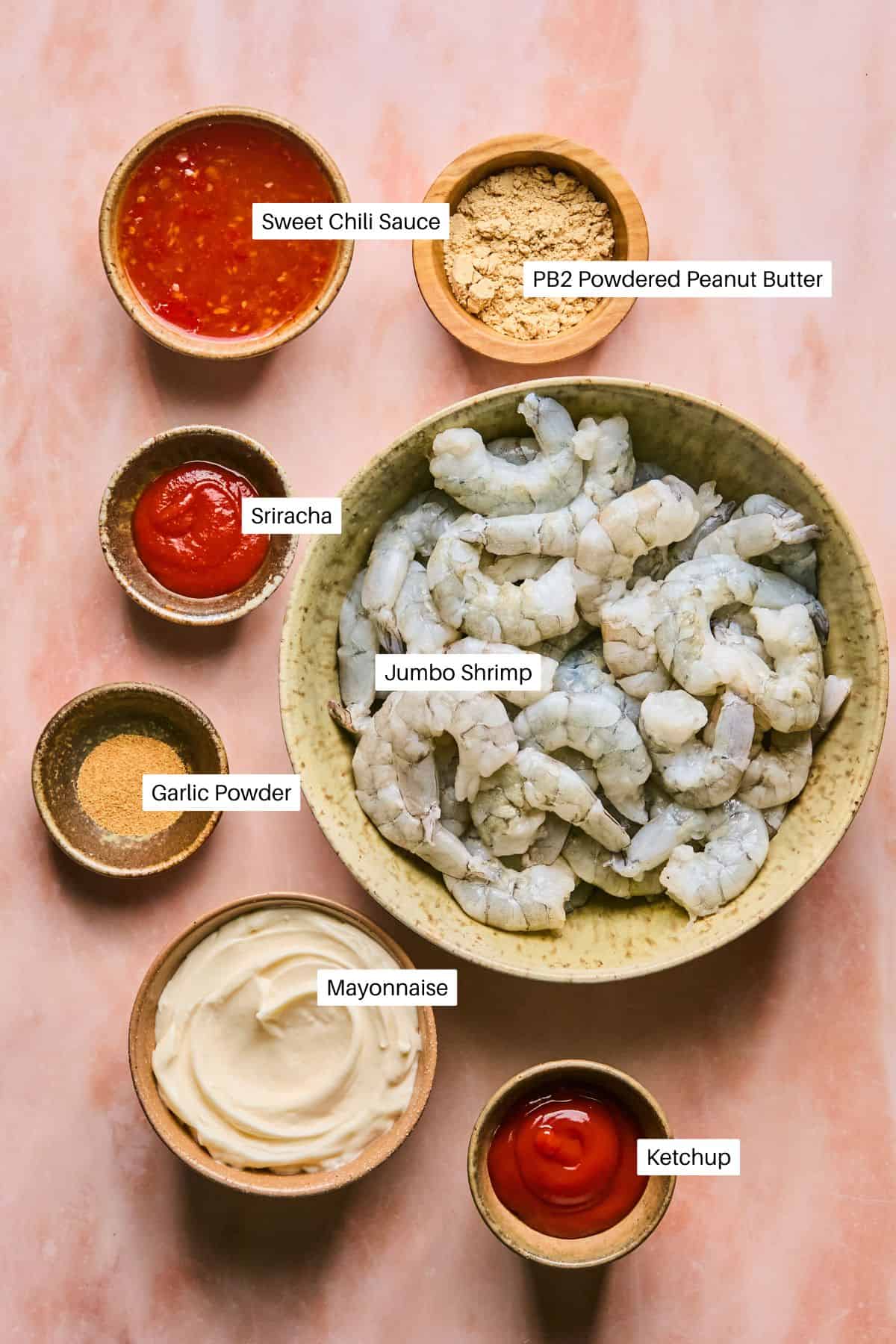 The image showcases ingredients for a boom boom shrimp recipe: a bowl of jumbo shrimp, bowls of sweet chili sauce, powdered peanut butter, sriracha, garlic powder, mayonnaise, and ketchup. Each item is labeled on the light pink surface.