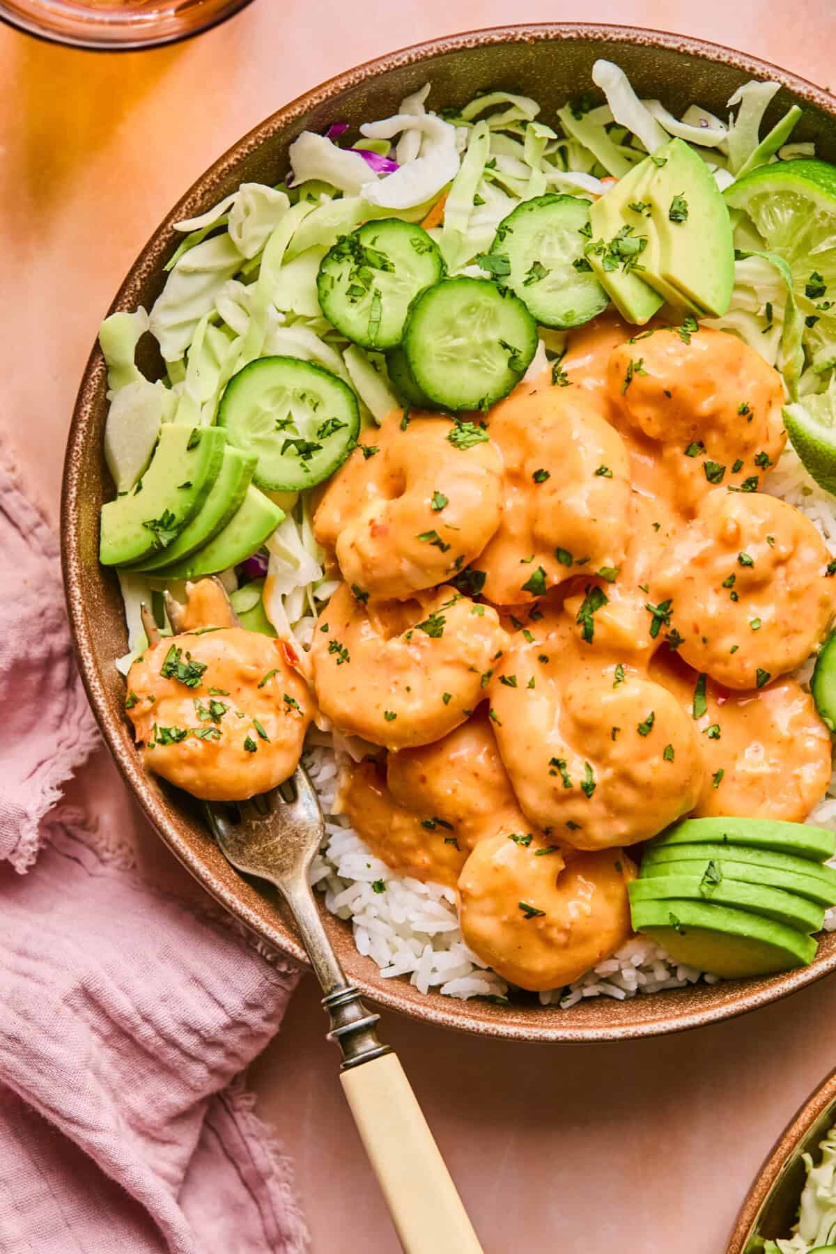 A bowl of creamy boom boom shrimp with a vibrant orange sauce is served over white rice. Its garnished with sliced avocado, cucumber, and shredded cabbage. A fork rests on the side, and a pink cloth is partially visible at the bottom.