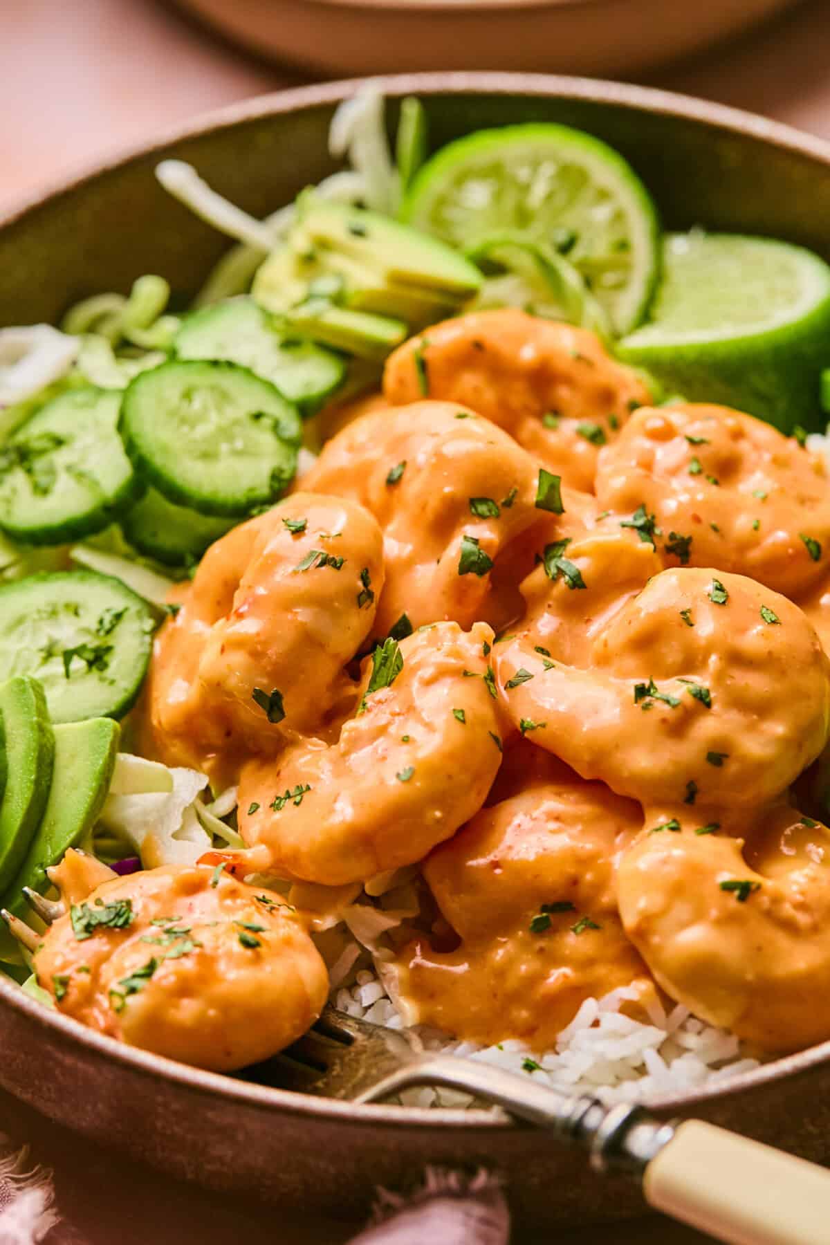 A bowl of creamy boom boom shrimp with white rice, garnished with slices of avocado, cucumber, and lime. The shrimp is coated in an orange sauce and sprinkled with herbs. A fork rests in the bowl.