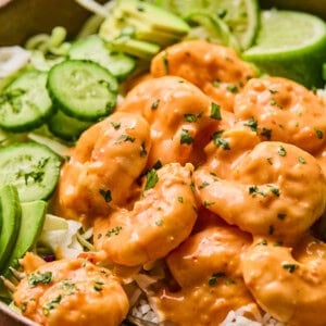 A bowl of creamy boom boom shrimp with white rice, garnished with slices of avocado, cucumber, and lime. The shrimp is coated in an orange sauce and sprinkled with herbs. A fork rests in the bowl.