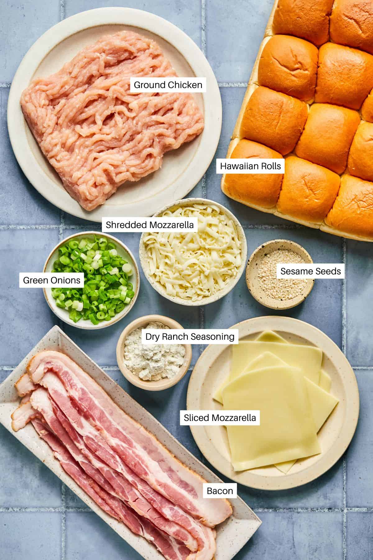 Top view of labeled ingredients for chicken bacon ranch sliders on a tiled surface: ground chicken, Hawaiian rolls, green onions, shredded mozzarella, sesame seeds, dry ranch seasoning, sliced mozzarella, and crispy bacon.