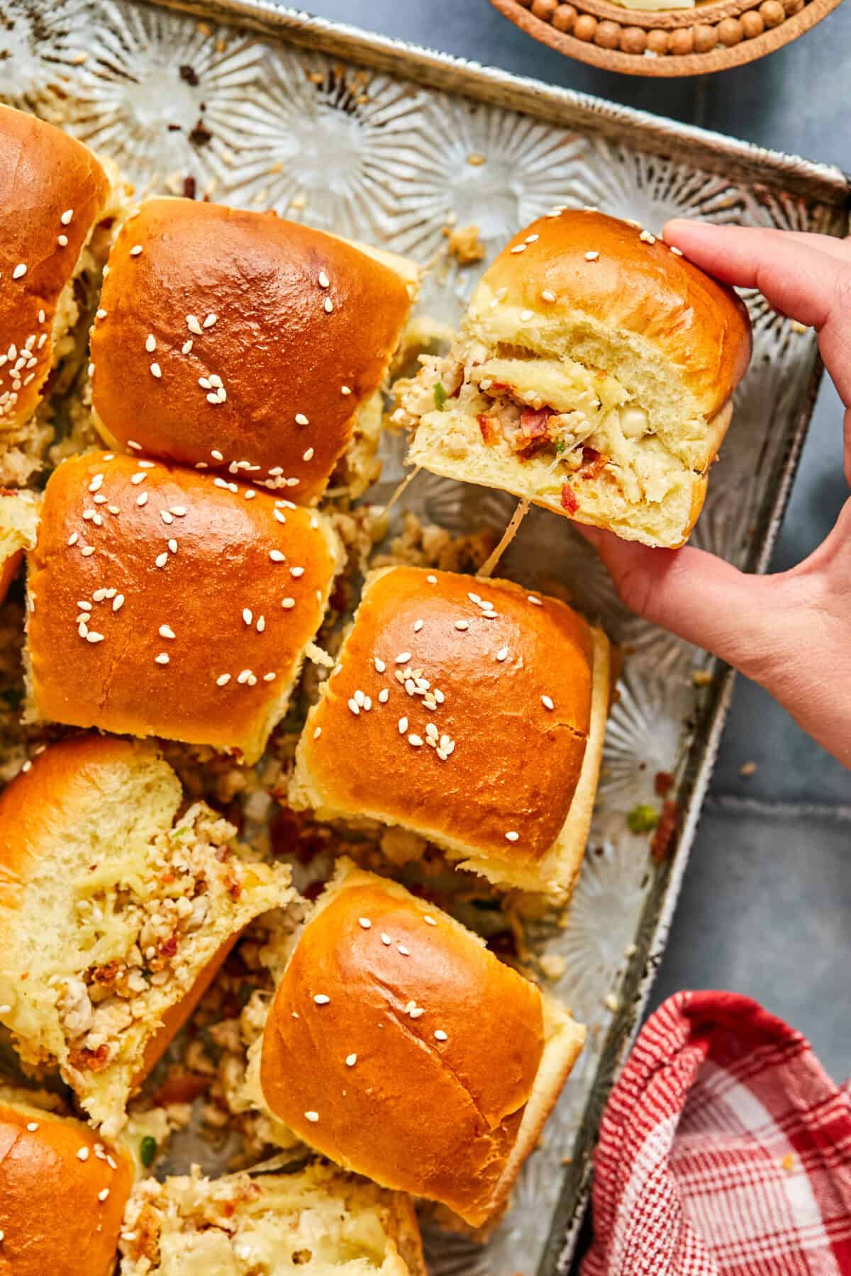 A tray of golden-brown chicken bacon ranch sliders with sesame seed tops awaits. One slider is being picked up, unveiling a luscious filling of melted cheese, diced vegetables, and savory goodness. A red and white checkered cloth adds a charming touch on the side.