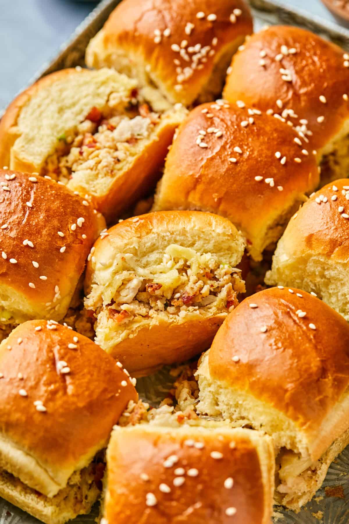 A close-up of freshly baked, fluffy chicken bacon ranch sliders filled with a savory mixture, topped with sesame seeds. The golden-brown tops glisten, suggesting a soft, buttery texture. The filling includes finely chopped ingredients, peeking out from the split rolls.