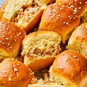 A close-up of freshly baked, fluffy chicken bacon ranch sliders filled with a savory mixture, topped with sesame seeds. The golden-brown tops glisten, suggesting a soft, buttery texture. The filling includes finely chopped ingredients, peeking out from the split rolls.