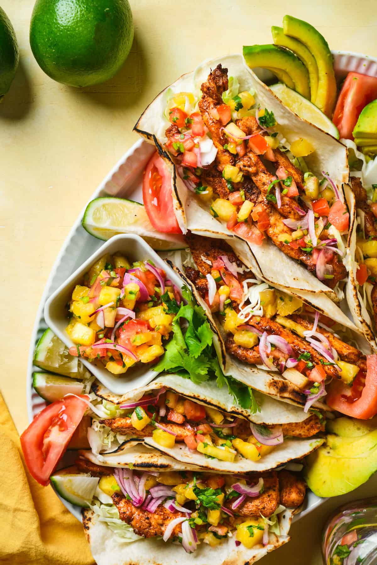 A plate of blackened chicken tacos filled with diced tomatoes, onions, and fresh cilantro. The tacos are served with lime wedges and garnished with avocado slices. Two bowls of pineapple salsa and slices of limes are beside the plate.