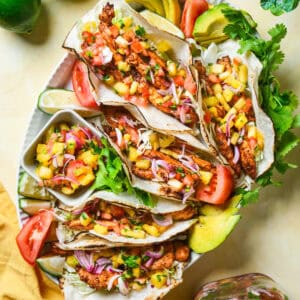 A platter of blackened chicken tacos filled with diced pineapple, red onions, and cilantro. Sliced avocados and tomatoes surround the tacos. Fresh limes and a jar of salsa are nearby. A yellow napkin and green leaves are visible in the background.