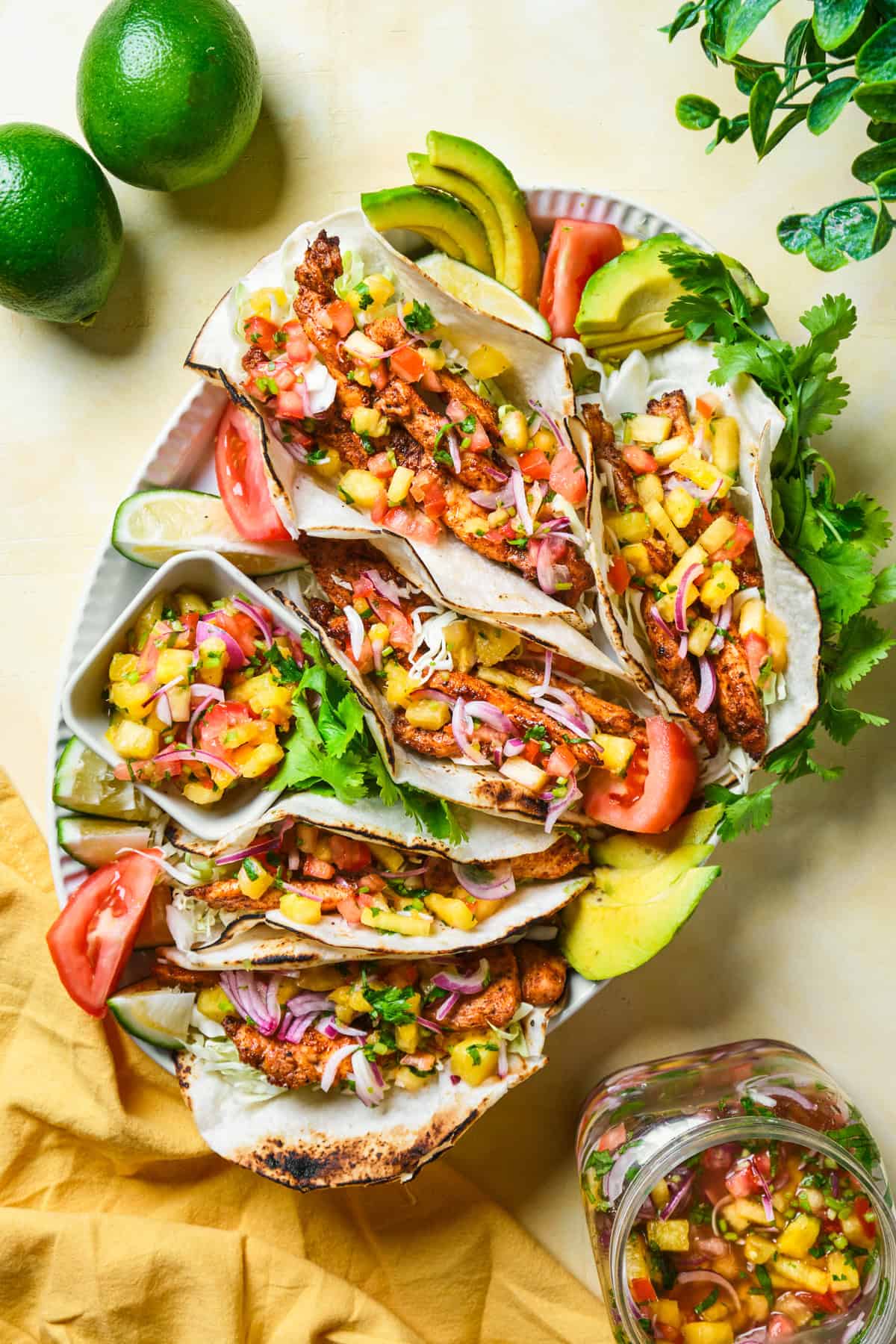 A platter of blackened chicken tacos filled with diced pineapple, red onions, and cilantro. Sliced avocados and tomatoes surround the tacos. Fresh limes and a jar of salsa are nearby. A yellow napkin and green leaves are visible in the background.