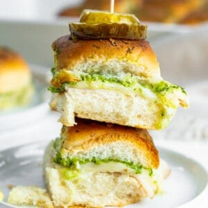 These Chicken Pesto Sliders are stacked mini sandwiches with turkey, cheese, and pesto on soft rolls. Each slider is topped with pickle slices and secured with a toothpick, elegantly arranged on a white plate against a bright background.