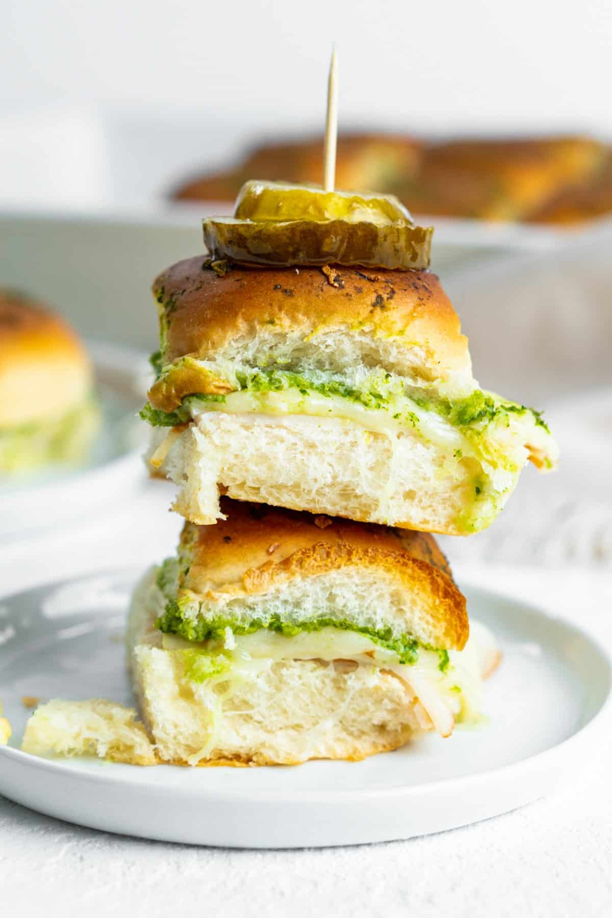 These Chicken Pesto Sliders are stacked mini sandwiches with turkey, cheese, and pesto on soft rolls. Each slider is topped with pickle slices and secured with a toothpick, elegantly arranged on a white plate against a bright background.