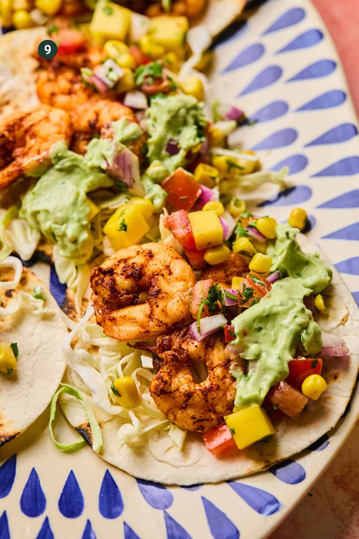 Close-up of air fryer shrimp tacos with vibrant toppings on a decorative plate. The tacos are garnished with diced mango, corn, tomatoes, sliced red onions, cilantro, and a dollop of creamy green sauce on shredded cabbage, all on soft tortillas.