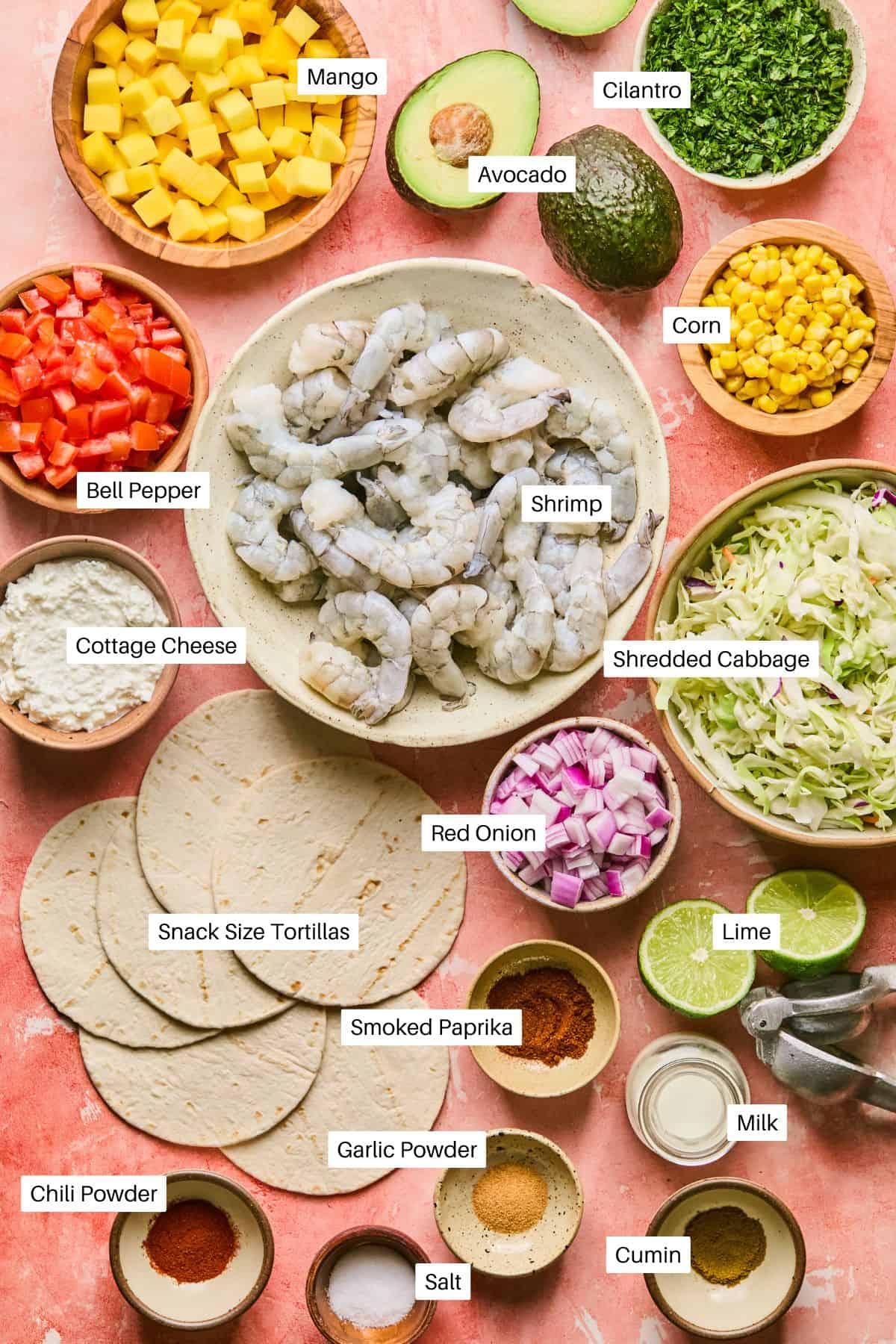 Ingredients for air fryer shrimp tacos on a pink surface include: chopped mango, cilantro, half an avocado, corn, bell pepper, shrimp, cottage cheese, shredded cabbage, red onion, snack size tortillas, lime, smoked paprika, chili powder, garlic powder, salt, cumin, and milk.