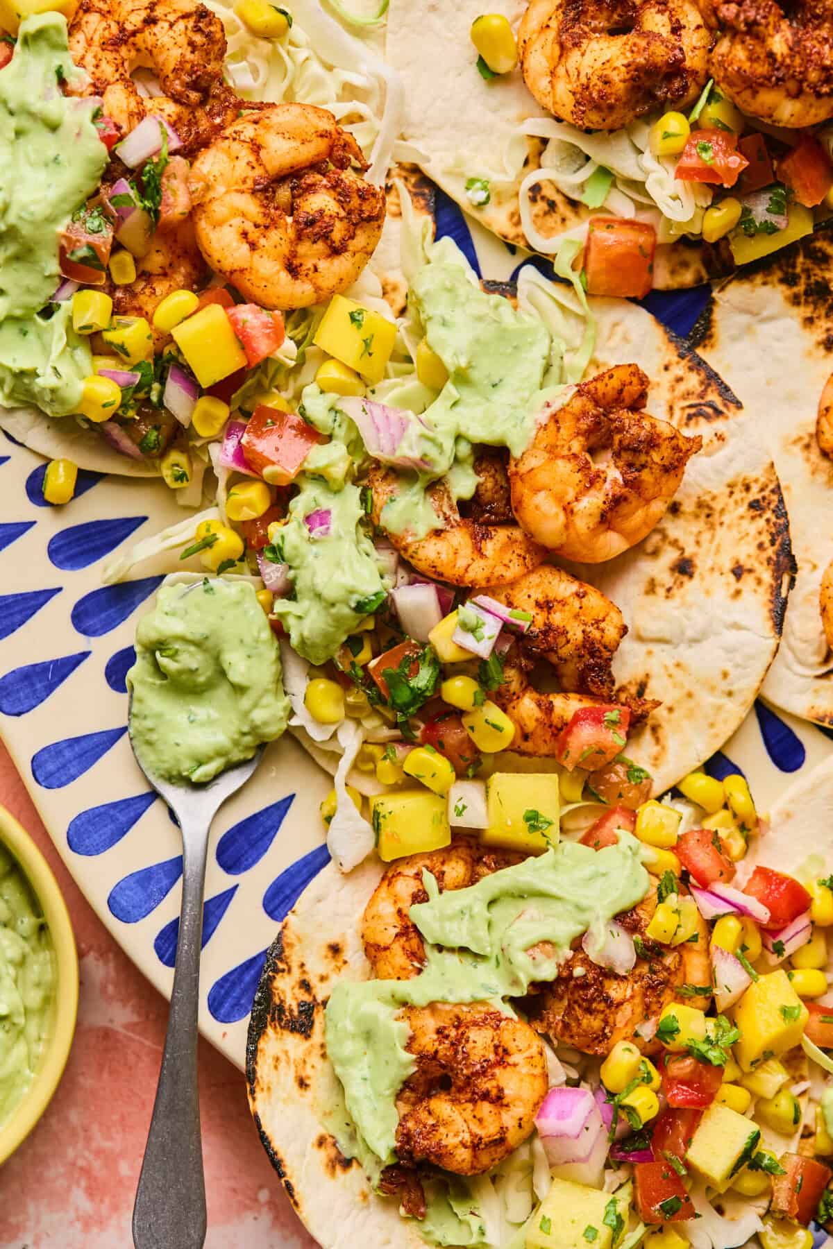 Air fryer shrimp tacos filled with grilled shrimp, diced mango, tomato, onion, and corn are drizzled with creamy avocado sauce. Theyre beautifully arranged on a decorative blue and white platter with a spoon of sauce resting beside them.