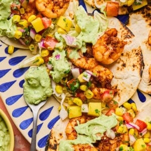 Air fryer shrimp tacos filled with grilled shrimp, diced mango, tomato, onion, and corn are drizzled with creamy avocado sauce. Theyre beautifully arranged on a decorative blue and white platter with a spoon of sauce resting beside them.