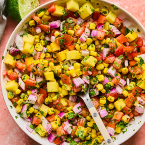 A vibrant bowl of mango corn salsa featuring diced mango, tomatoes, red onion, and fresh herbs. A halved lime sits beside the bowl on a textured pink surface, with a spoon nestled in the delicious mix.