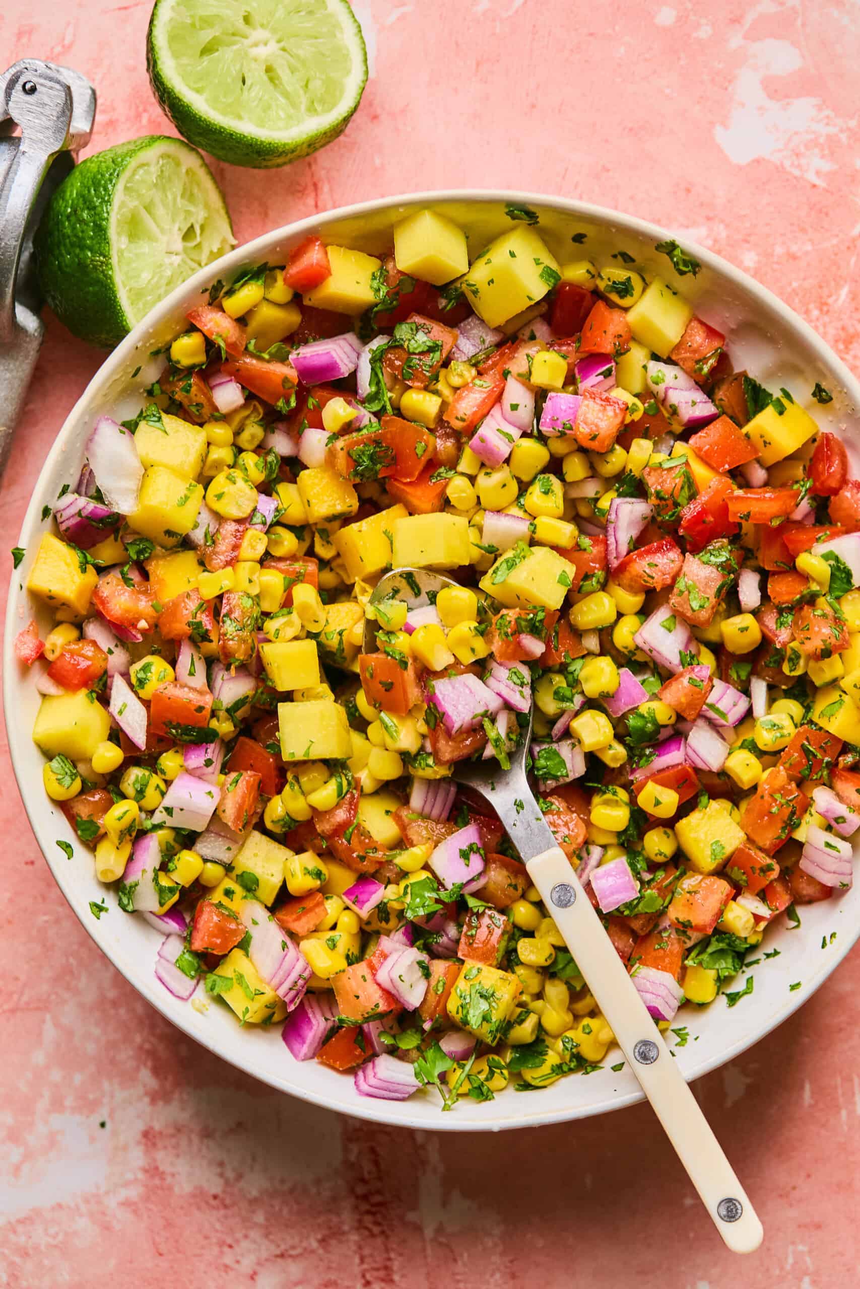 A vibrant bowl of mango corn salsa featuring diced mango, tomatoes, red onion, and fresh herbs. A halved lime sits beside the bowl on a textured pink surface, with a spoon nestled in the delicious mix.