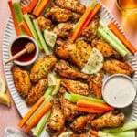 A large oval platter with Hot Honey Lemon Pepper Wings, celery sticks, carrot sticks, lemon slices, and bowls of ranch and honey dipping sauces sits on a pink table with two drinks, a bottle, and a striped napkin nearby.