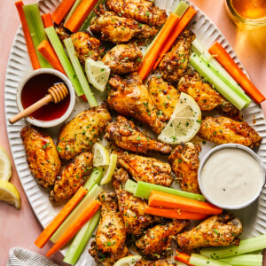 A large oval platter with Hot Honey Lemon Pepper Wings, celery sticks, carrot sticks, lemon slices, and bowls of ranch and honey dipping sauces sits on a pink table with two drinks, a bottle, and a striped napkin nearby.