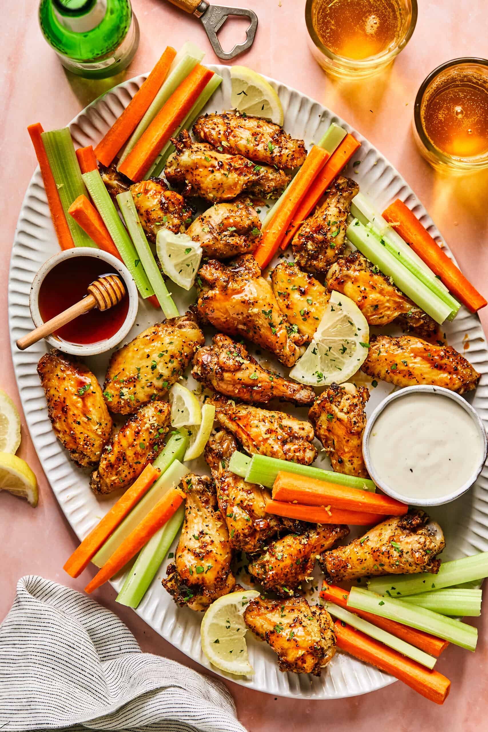 A large oval platter with Hot Honey Lemon Pepper Wings, celery sticks, carrot sticks, lemon slices, and bowls of ranch and honey dipping sauces sits on a pink table with two drinks, a bottle, and a striped napkin nearby.