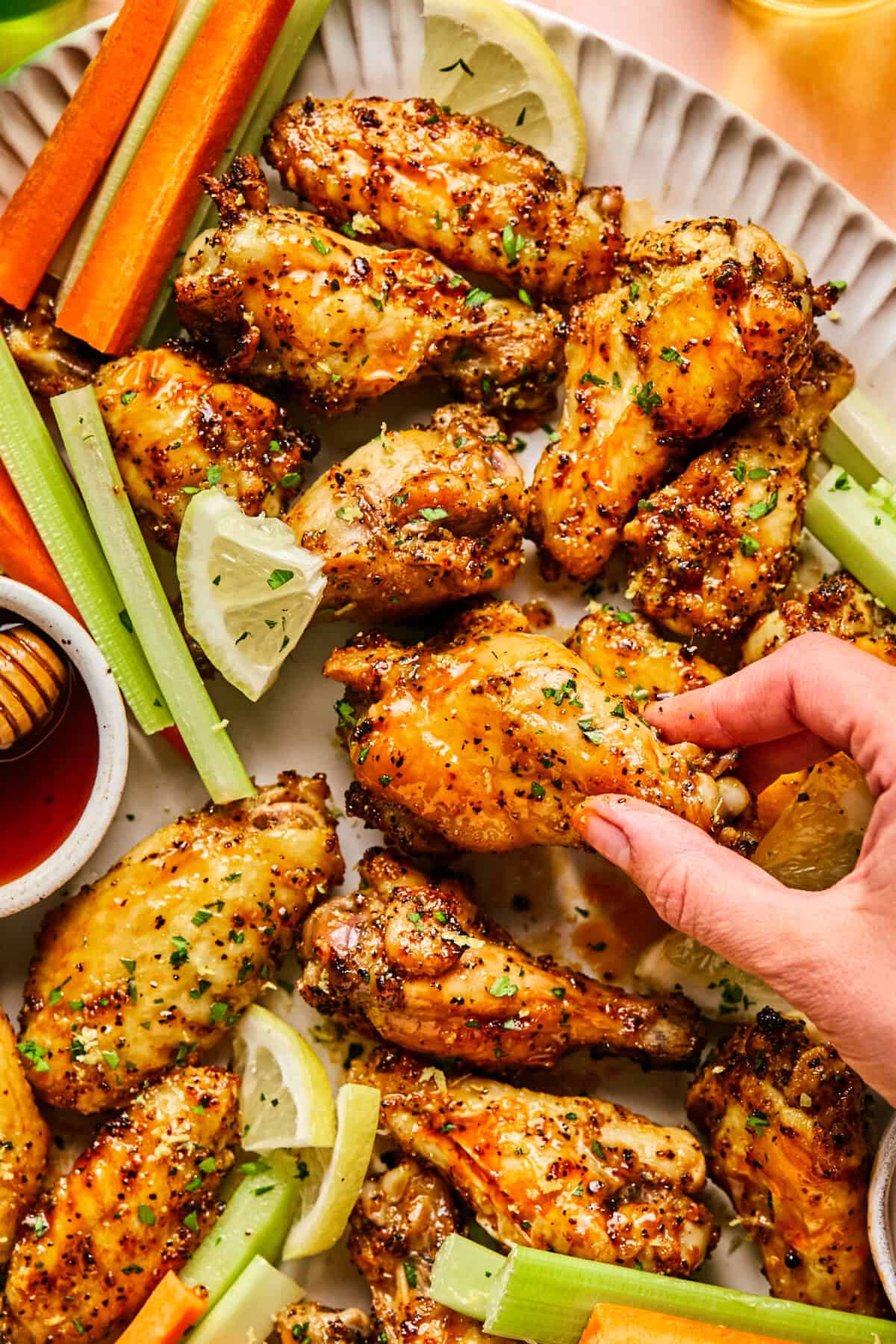 A hand reaches for a Hot Honey Lemon Pepper Wing on a platter served with celery sticks, carrot sticks, lemon wedges, and ramekins of honey and dipping sauce.