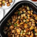 Air Fryer Breakfast Potatoes: Diced potatoes seasoned with herbs and garnished with chopped green onions in a black tray. Nearby are small white bowls with ketchup and extra toppings, alongside a plate teeming with more of these air-fried delights.