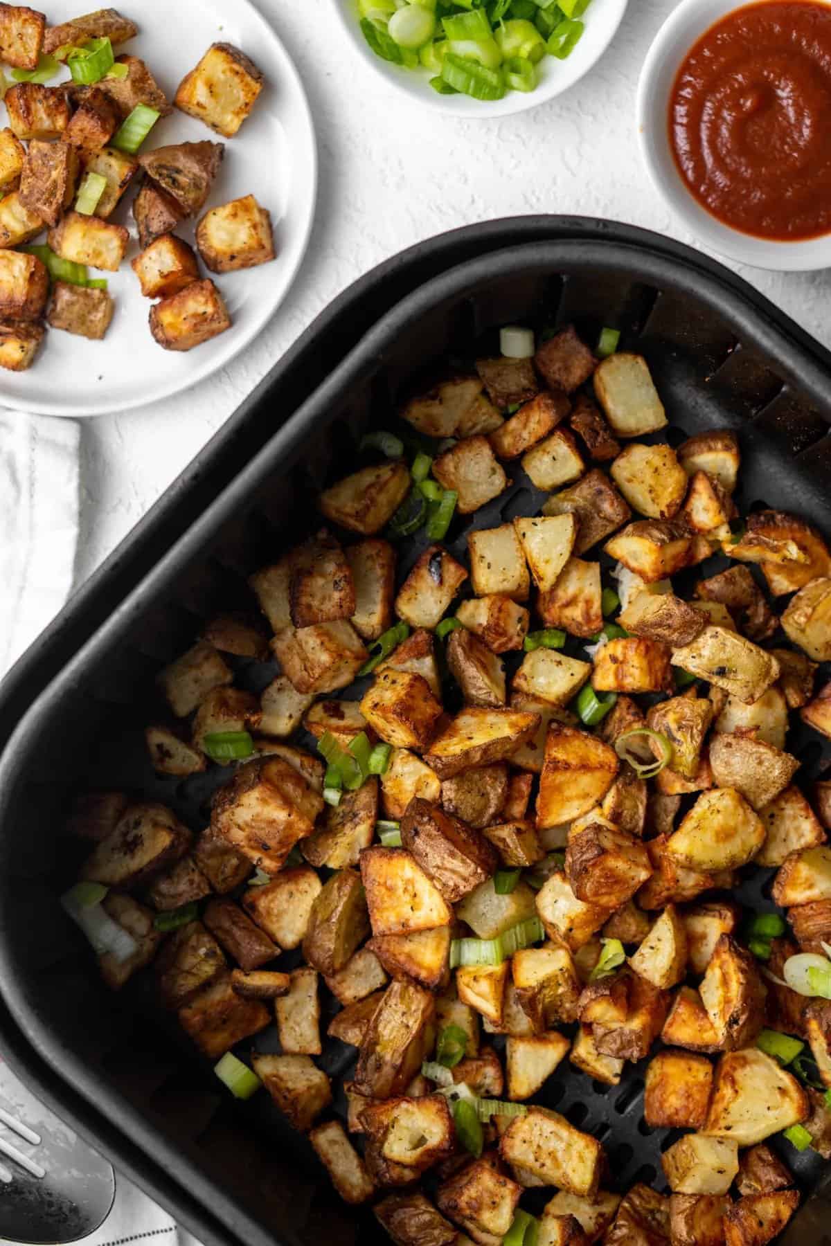 Air Fryer Breakfast Potatoes: Diced potatoes seasoned with herbs and garnished with chopped green onions in a black tray. Nearby are small white bowls with ketchup and extra toppings, alongside a plate teeming with more of these air-fried delights.