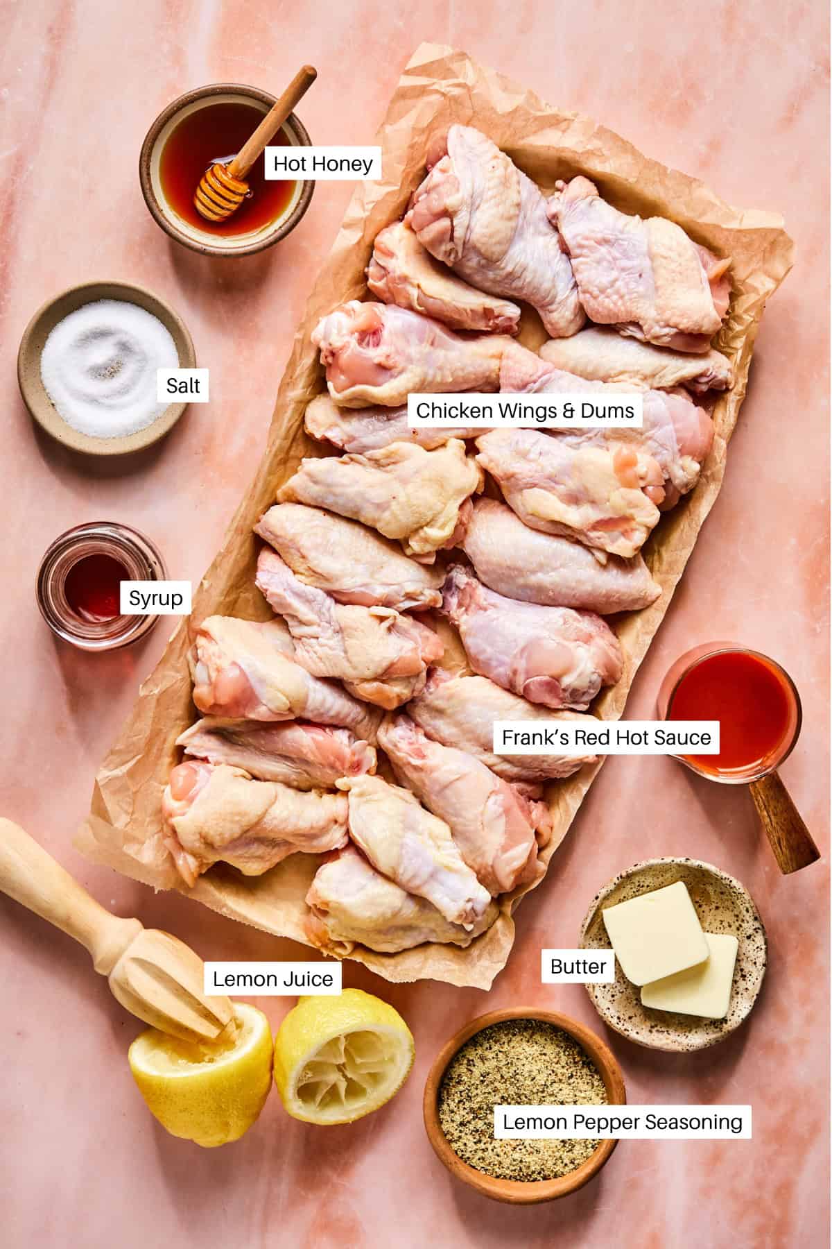 A tray of raw chicken wings, prepped for Hot Honey Lemon Pepper Wings, is surrounded by labeled ingredients—hot honey, salt, syrup, Frank’s Red Hot Sauce, lemon juice, butter, and lemon pepper seasoning—on a pink marble surface.