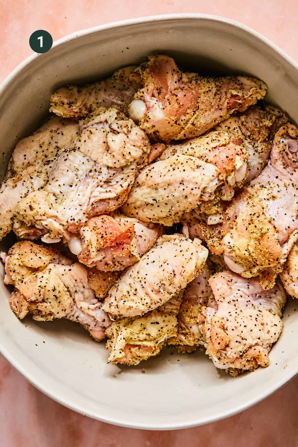 A large beige bowl filled with raw, seasoned chicken pieces coated in herbs and spices for Hot Honey Lemon Pepper Wings, set on a light pink surface. A small circle with the number 1 is in the top left corner of the image.
