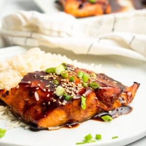 A plate features Air Fryer Teriyaki Salmon, glazed to perfection and garnished with sesame seeds and chopped green onions, served alongside fluffy white rice. A napkin is visible in the background.