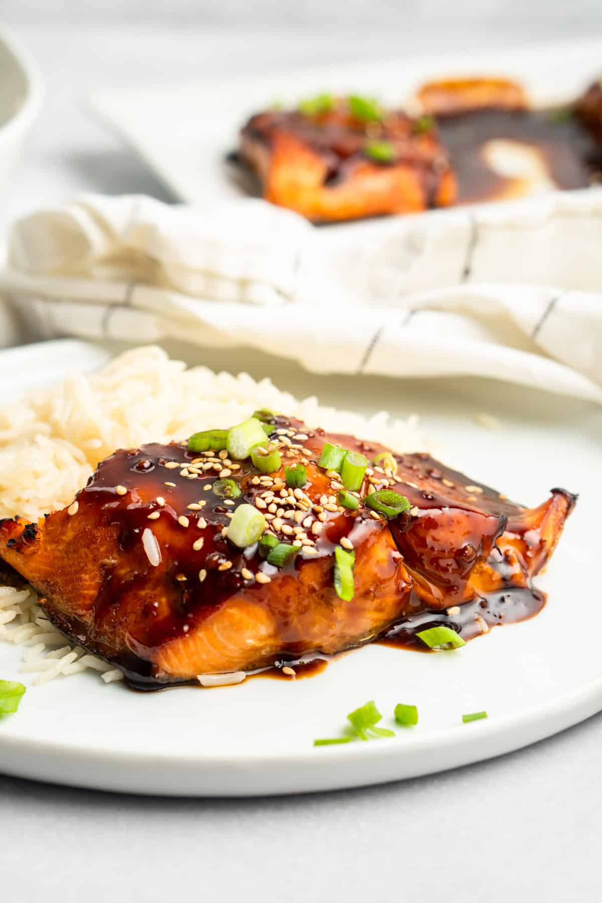 A plate features Air Fryer Teriyaki Salmon, glazed to perfection and garnished with sesame seeds and chopped green onions, served alongside fluffy white rice. A napkin is visible in the background.