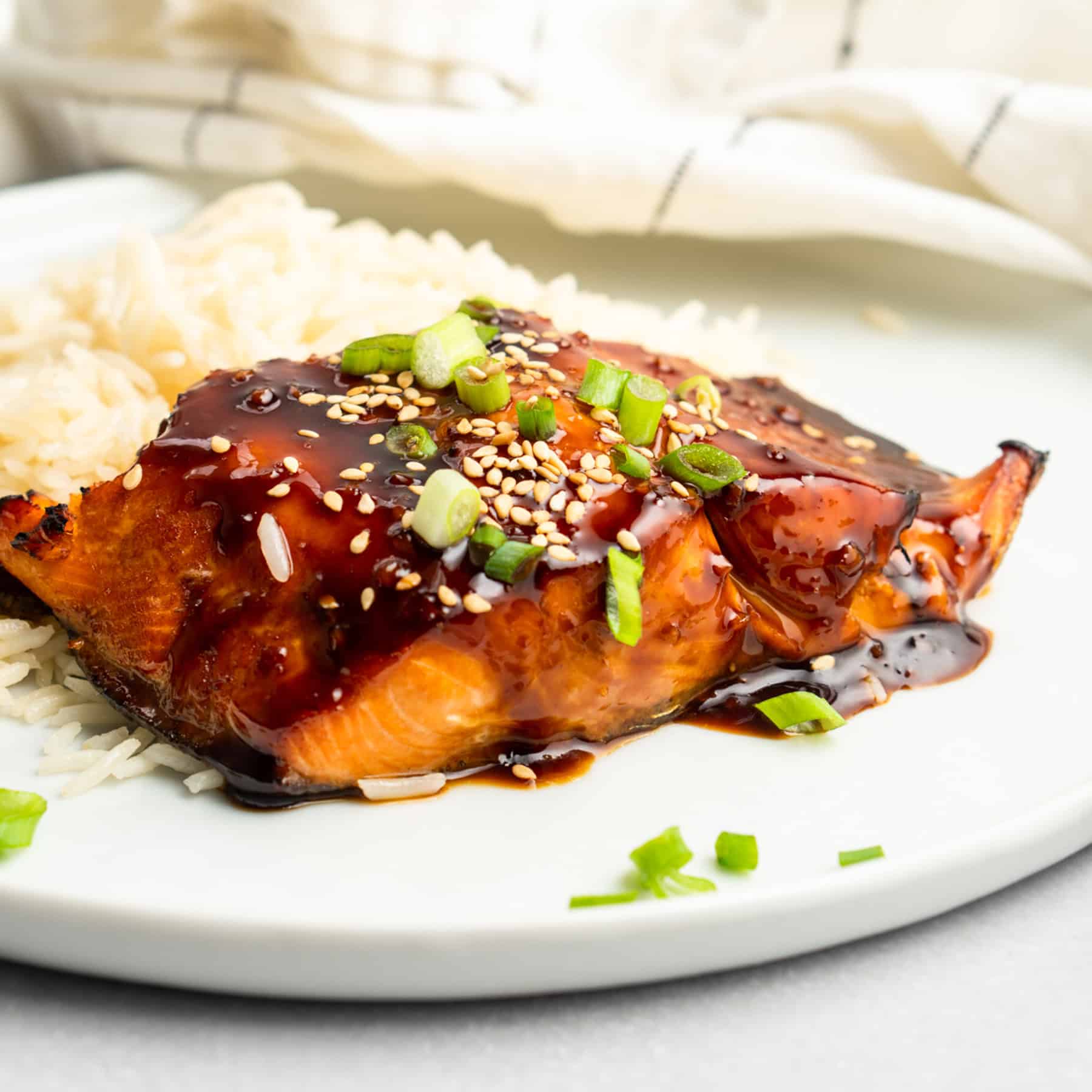 A plate featuring Air Fryer Teriyaki Salmon topped with sesame seeds and chopped green onions, served alongside a portion of white rice. A beige napkin is partially visible in the background.