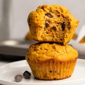 Two baked chocolate chip pumpkin muffins stacked on a plate with a bite taken out of the top muffin.