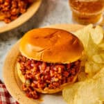 A Ground Chicken Sloppy Joe sandwich sits on a plate with potato chips. The toasted bun is filled with a rich, saucy meat mixture. A glass of soda and a checkered napkin nearby create a casual dining atmosphere.