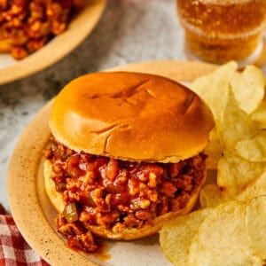 A Ground Chicken Sloppy Joe sandwich sits on a plate with potato chips. The toasted bun is filled with a rich, saucy meat mixture. A glass of soda and a checkered napkin nearby create a casual dining atmosphere.