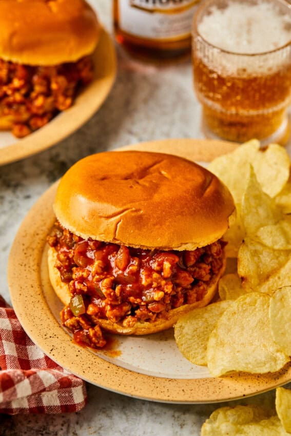 A Ground Chicken Sloppy Joe sandwich sits on a plate with potato chips. The toasted bun is filled with a rich, saucy meat mixture. A glass of soda and a checkered napkin nearby create a casual dining atmosphere.