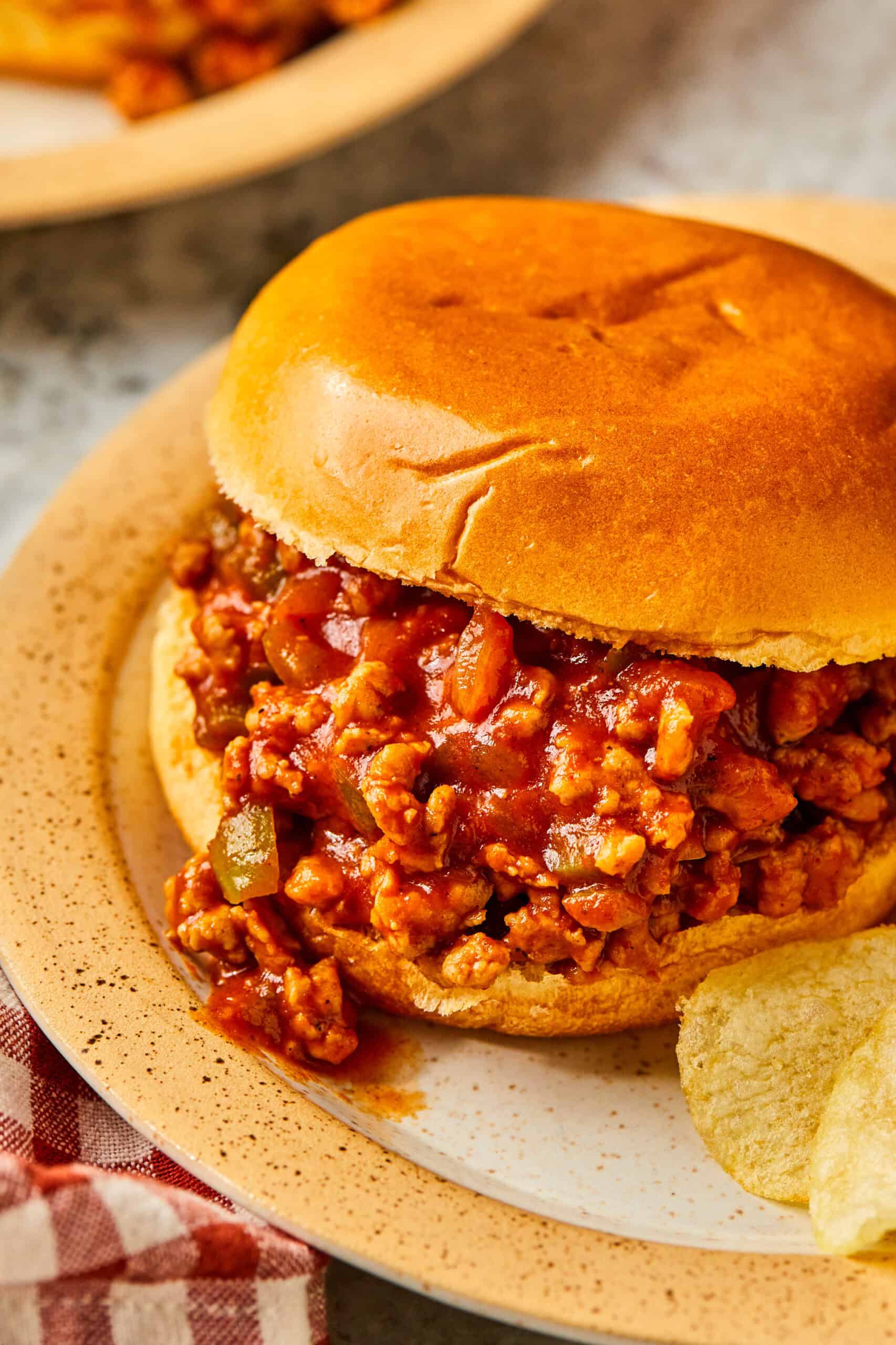 A close-up of a Ground Chicken Sloppy Joe sandwich on a beige plate. The sandwich is filled with a rich, tomato-based meat sauce and is accompanied by a few potato chips. The bun is golden brown, and a red checkered cloth is partially visible.