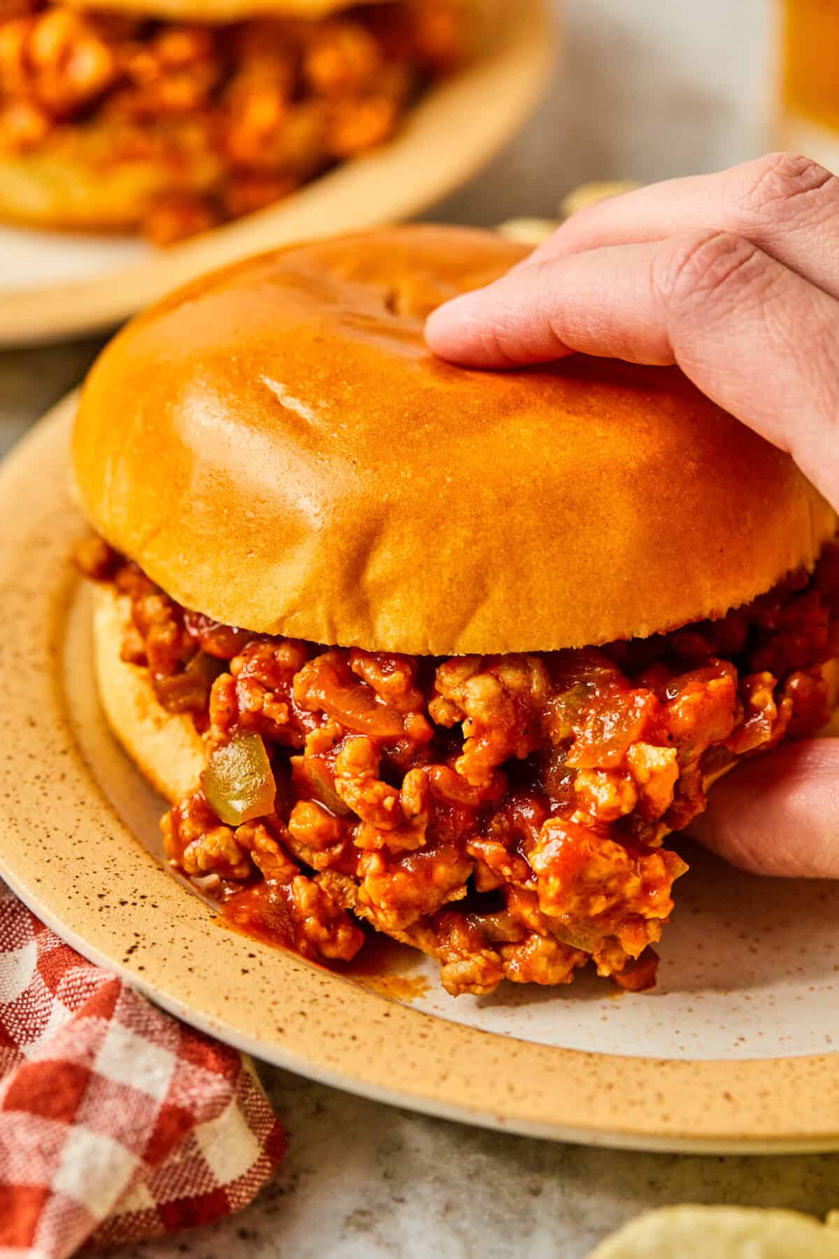 A close-up of a persons hand holding a Ground Chicken Sloppy Joe sandwich on a plate. The sandwich is filled with seasoned minced meat in a bun. A red checkered napkin is visible nearby.