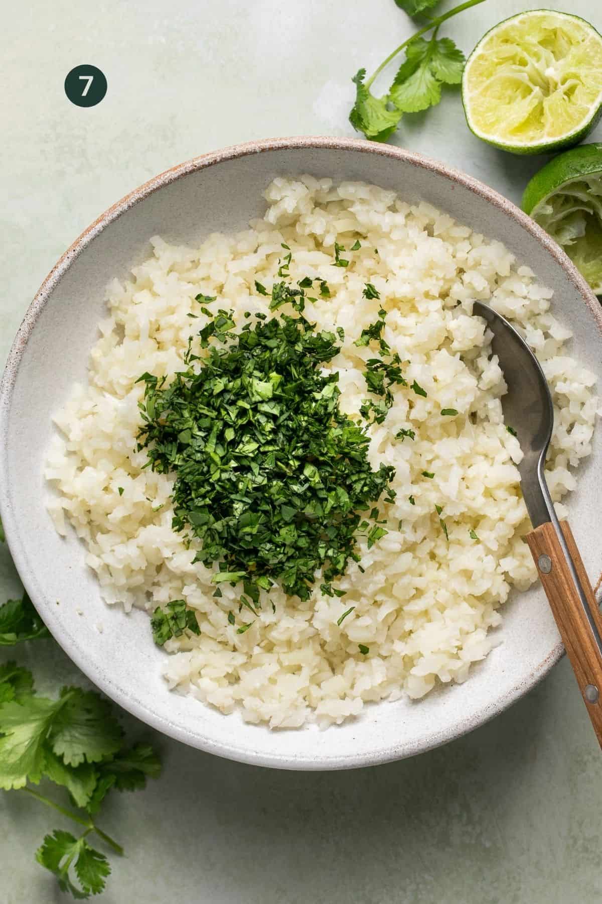 Cooked cauliflower rice, lime juice and cilantro added to a bowl to combine with a spoon.
