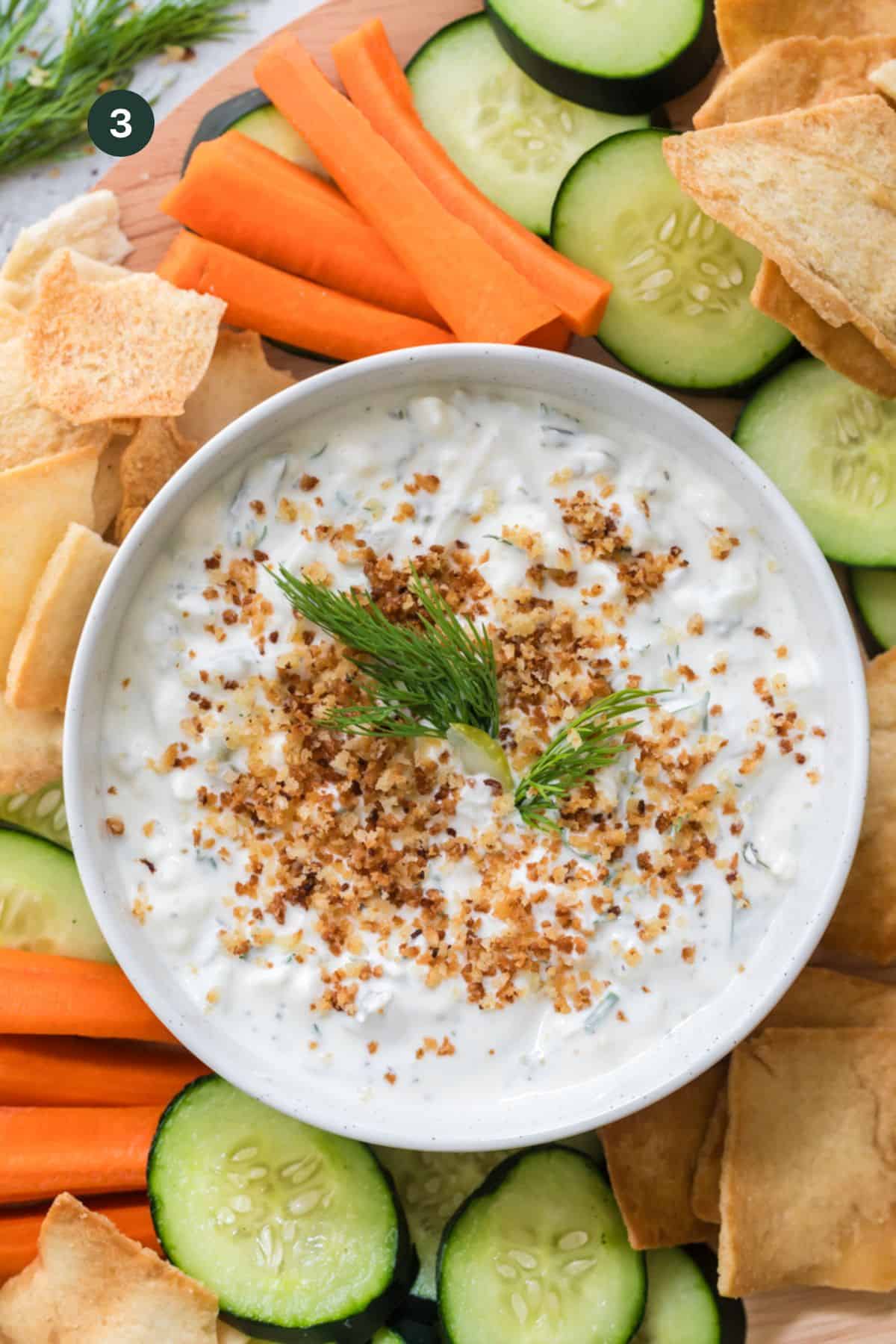 Dip topped with the remaining crunch panko and fresh dill in a bowl surrounded by carrots, cucumbers and pita chips.