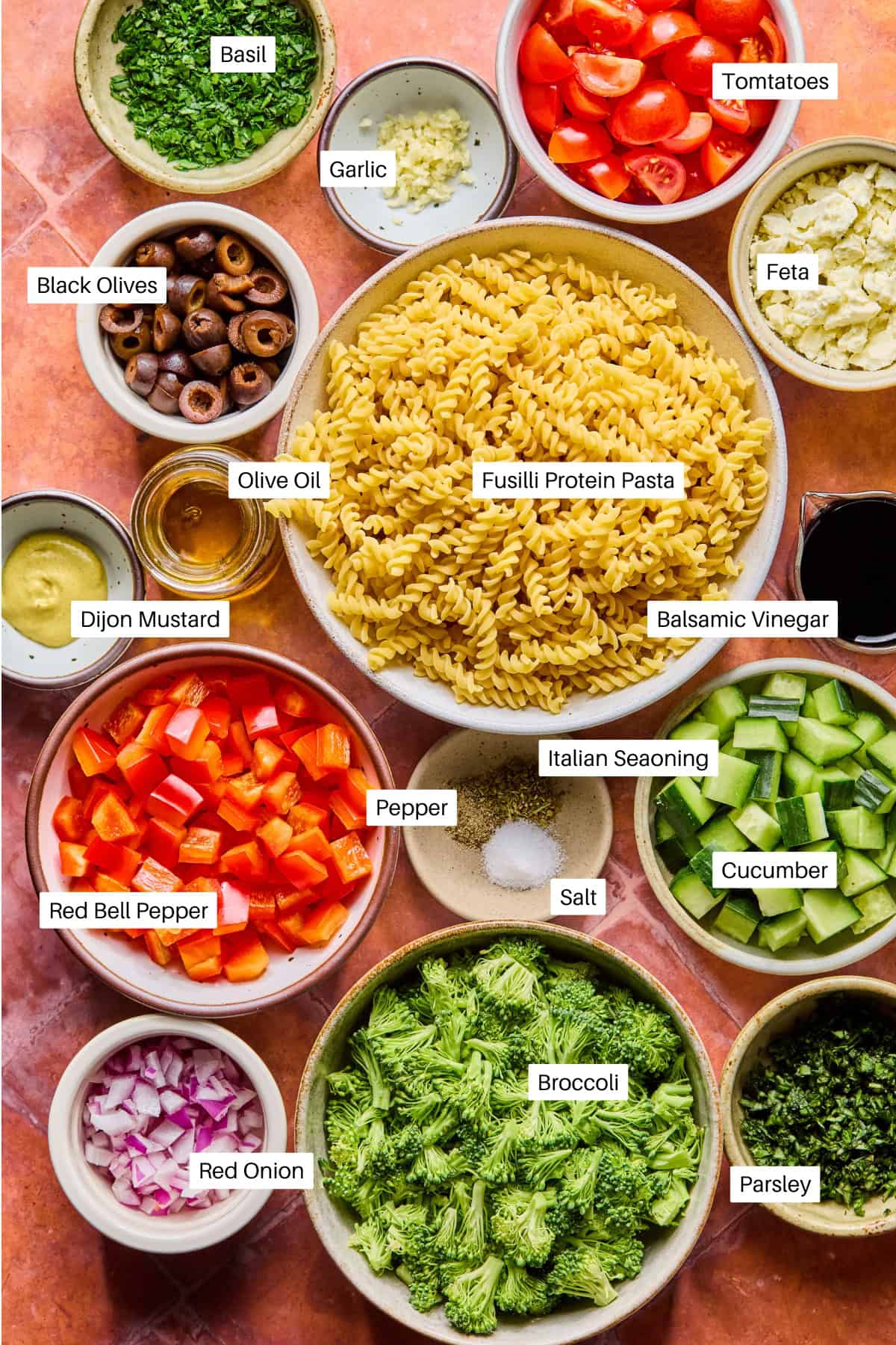 A variety of labeled ingredients for a high protein pasta salad, including fusilli protein pasta, tomatoes, feta, black olives, cucumber, broccoli, red bell pepper, red onion, olive oil, balsamic vinegar, Dijon mustard, spices, garlic, basil, and parsley.